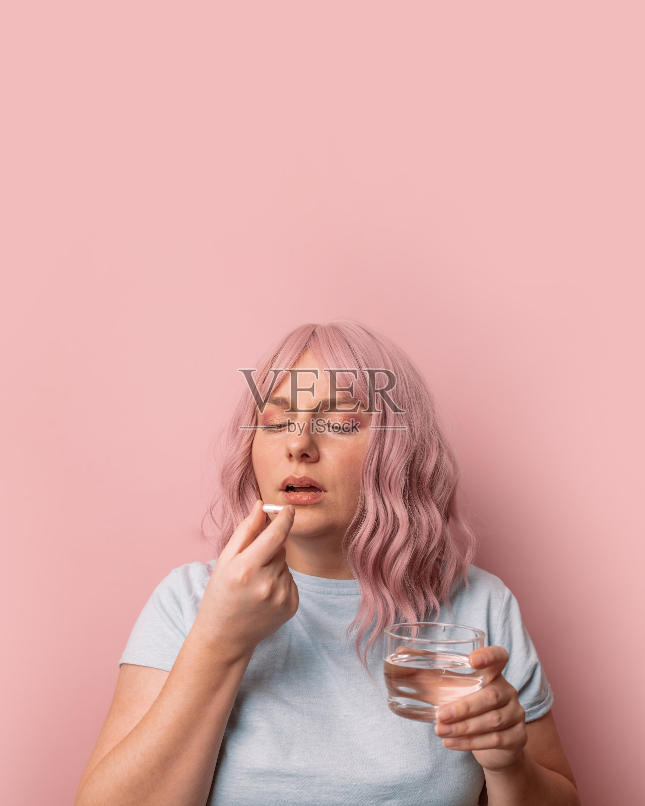 生病不舒服的年轻女子喝水，而遭受偏头痛孤立在粉红色的墙壁背景。卫生保健的概念照片摄影图片