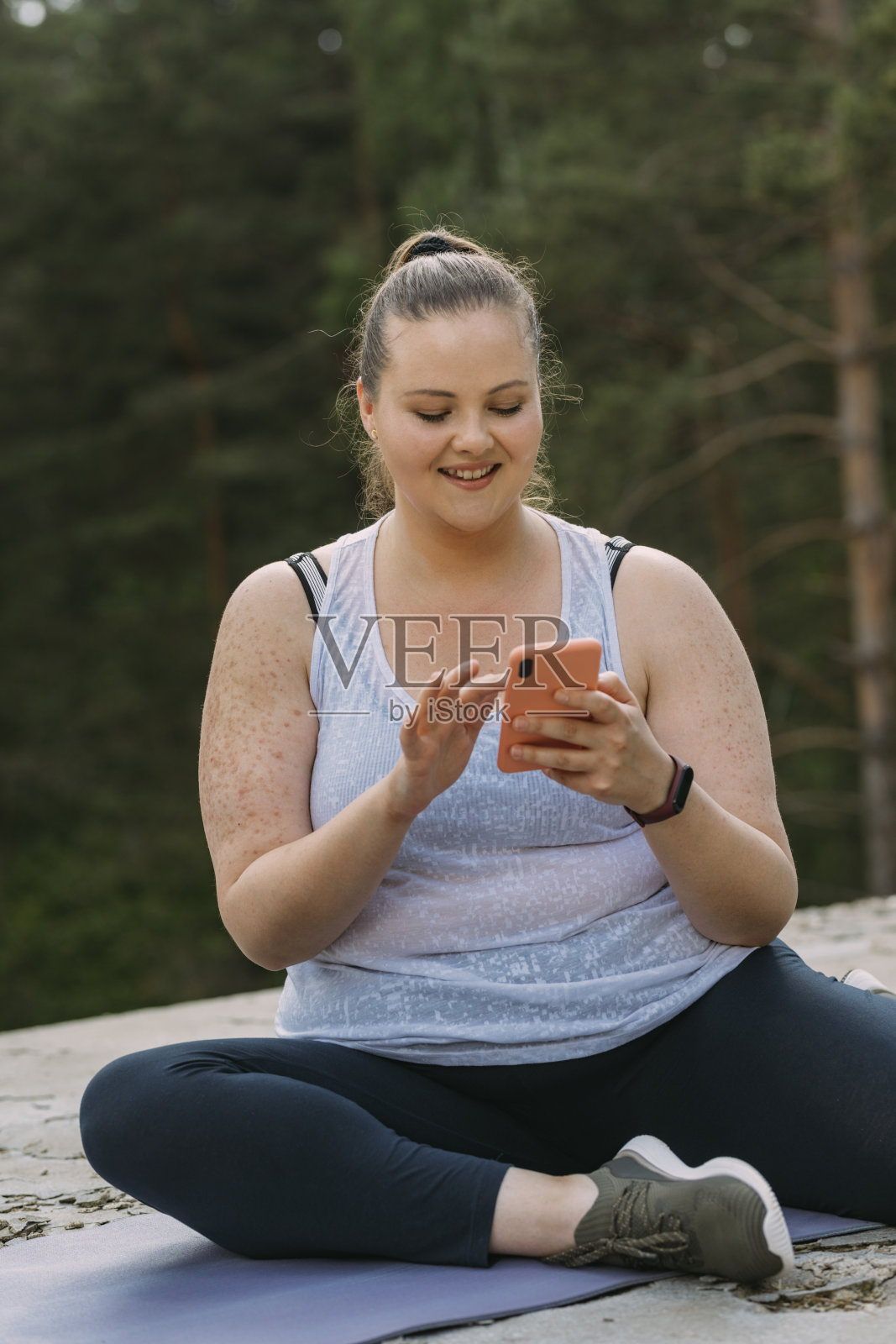 一个快乐的超重妇女坐在瑜伽垫上，在森林里锻炼后休息时用手机发短信照片摄影图片