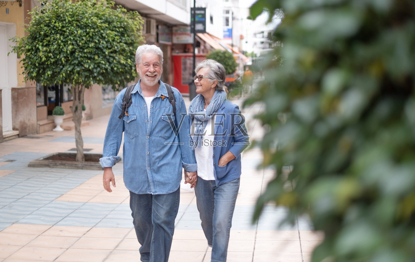 快乐、活跃、微笑的成年白种人老年夫妇手牵着手走在城市里作为游客。迷人的白发、穿着牛仔布的人们享受着自由、退休、度假和旅行照片摄影图片