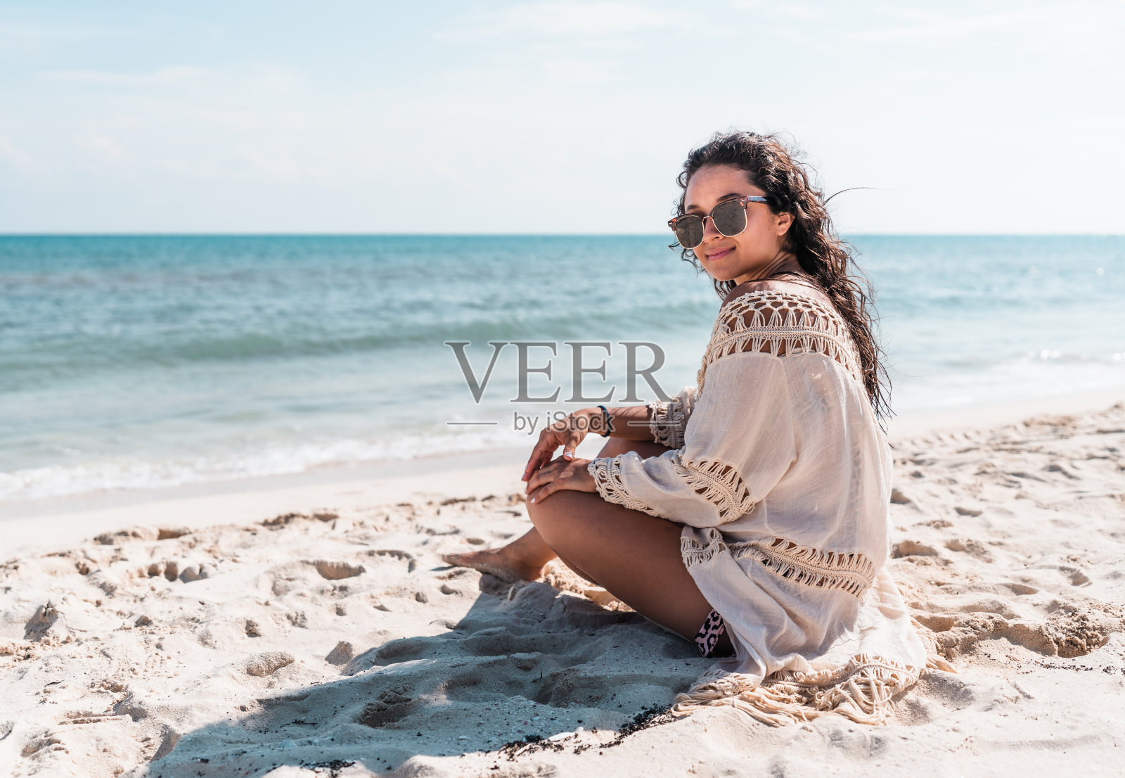 一位年轻的西班牙裔妇女，戴着太阳镜，穿着和服，微笑着坐在加勒比海海岸的沙滩上照片摄影图片