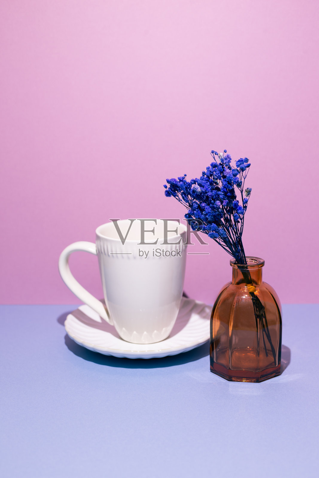 白色咖啡杯，蓝色的桌子上放着一瓶蓝色干花。粉红色的背景墙照片摄影图片