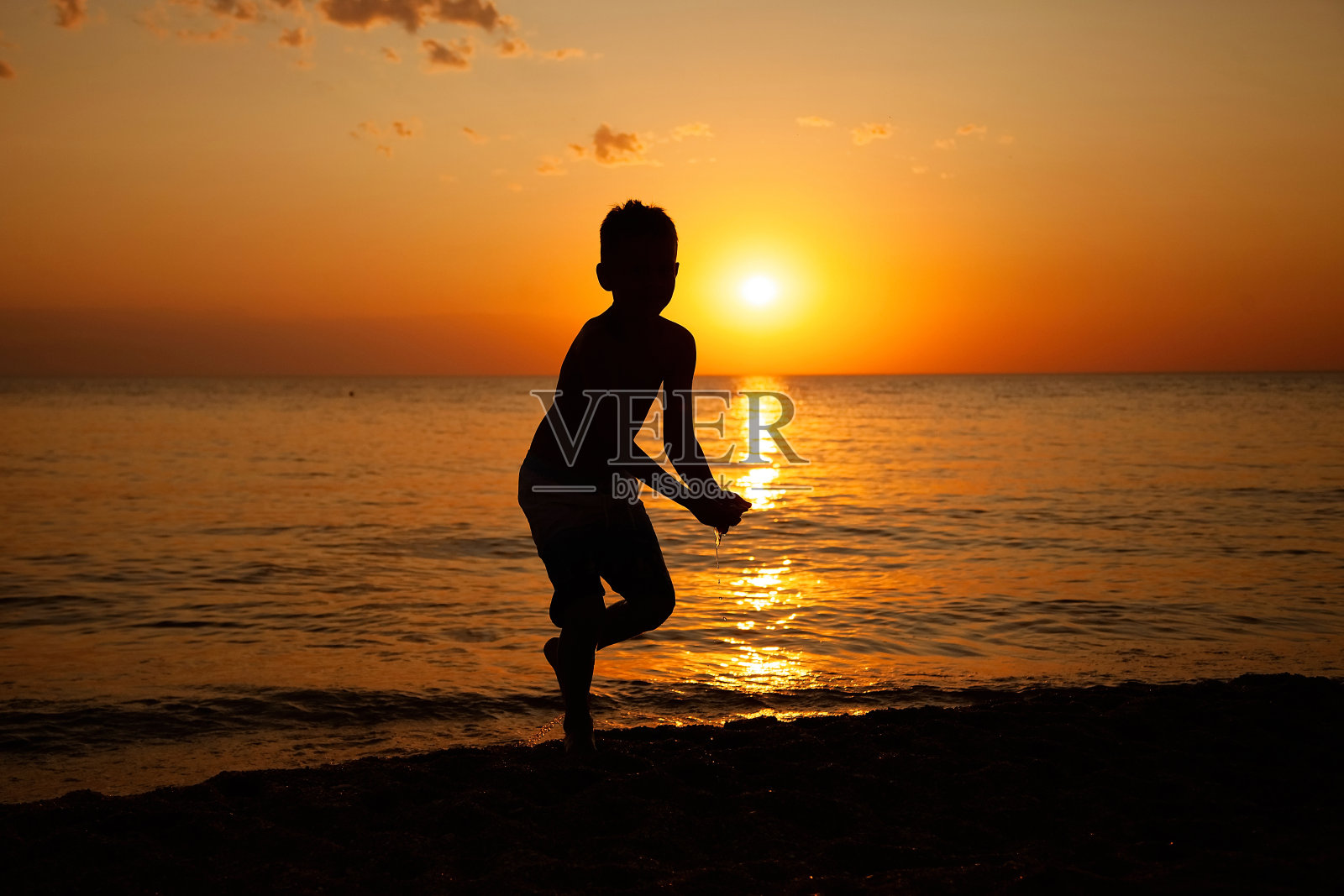 一个可爱的男孩从海里走出来，从他的手掌里拿出水，水从他的手在夏天的傍晚日落的海滩上滴下来照片摄影图片