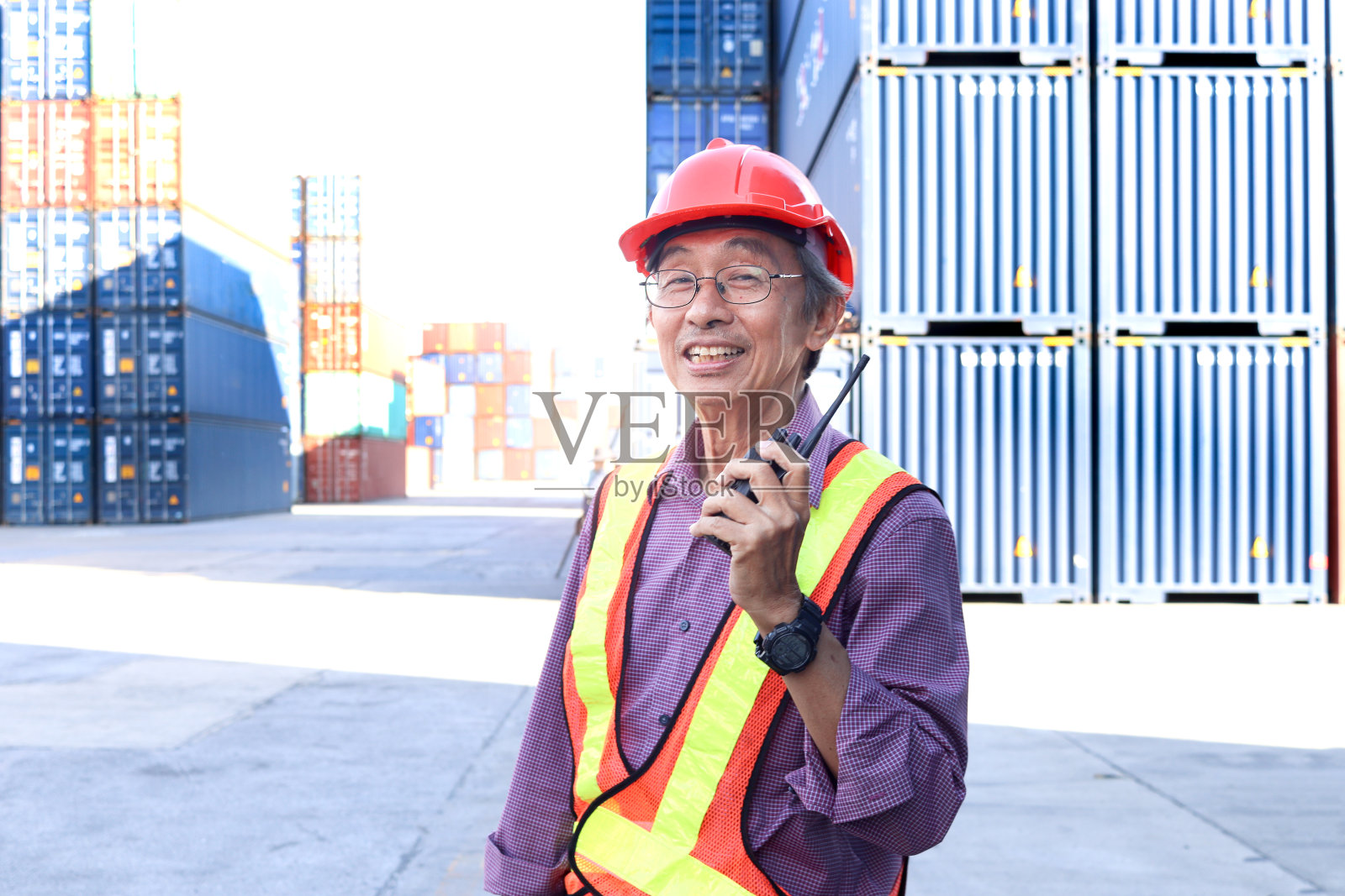 一位年长的亚洲工人工程师穿着安全背心和安全帽，站在物流运输货场，手持无线电对讲机。老年人在工作场所的概念。照片摄影图片