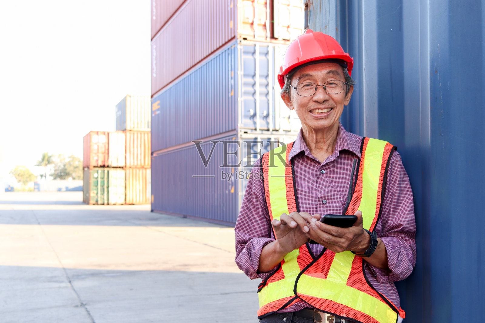 在物流运输集装箱堆场，一名年长的亚洲工人和工程师穿着安全背心和安全帽，站在那里用手机微笑。照片摄影图片