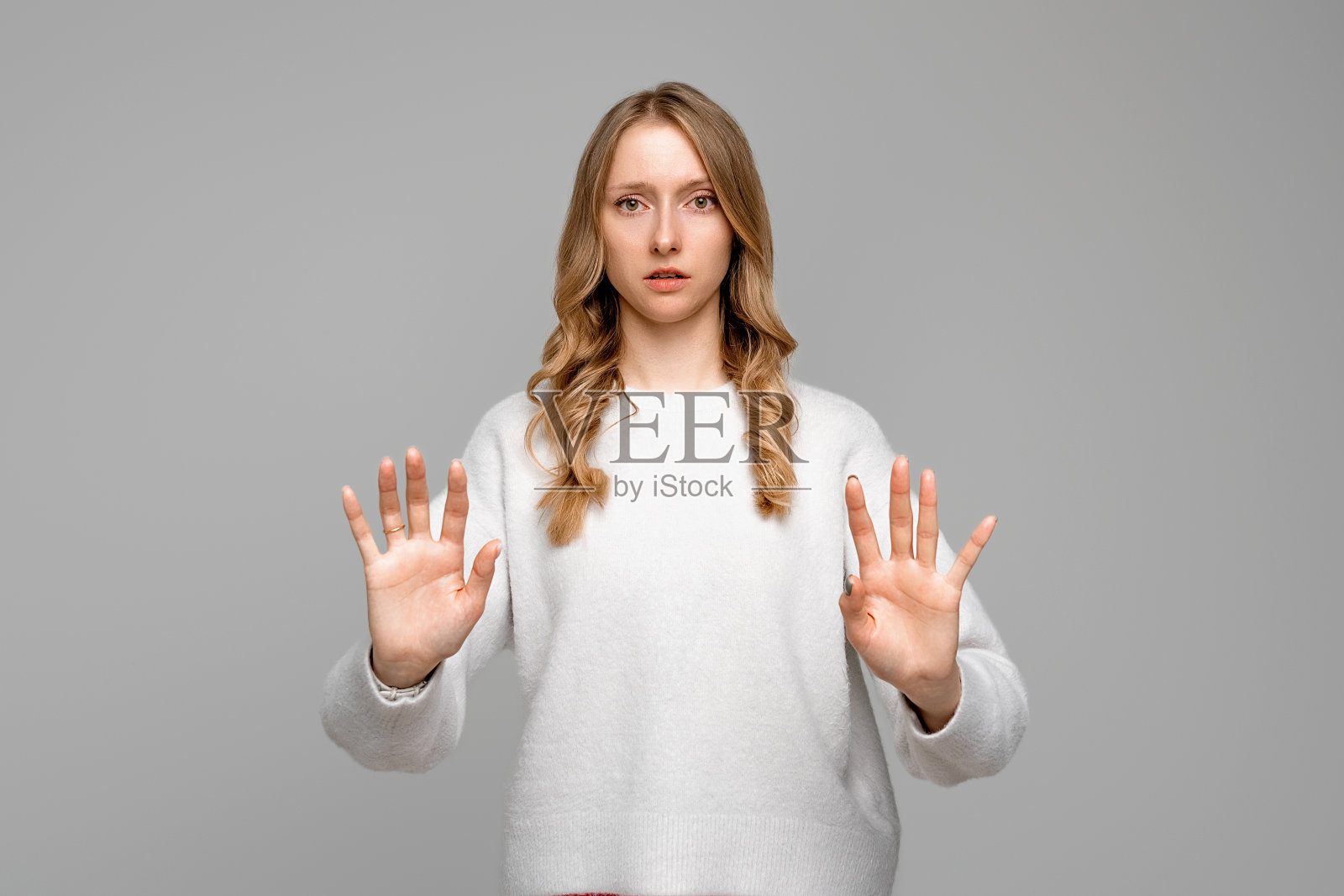 一个严肃的金发年轻女子，双手摆出停止的姿势，试图保护自己，好像在说:离我远点，穿白色毛衣，灰色背景。呆在家里。社会距离。新常态行为照片摄影图片