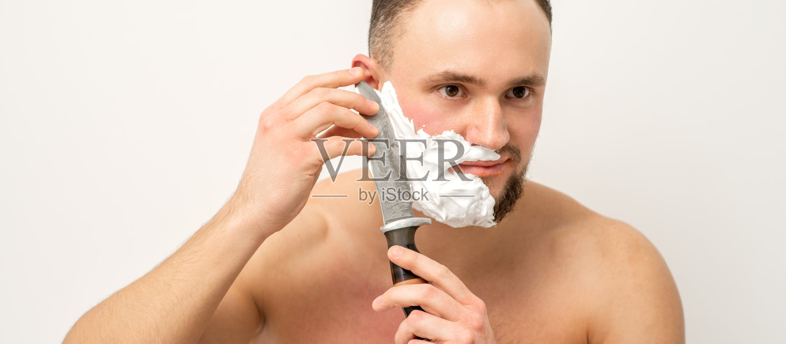 年轻的白人男子剃胡子用一把大刀在白色的背景。照片摄影图片