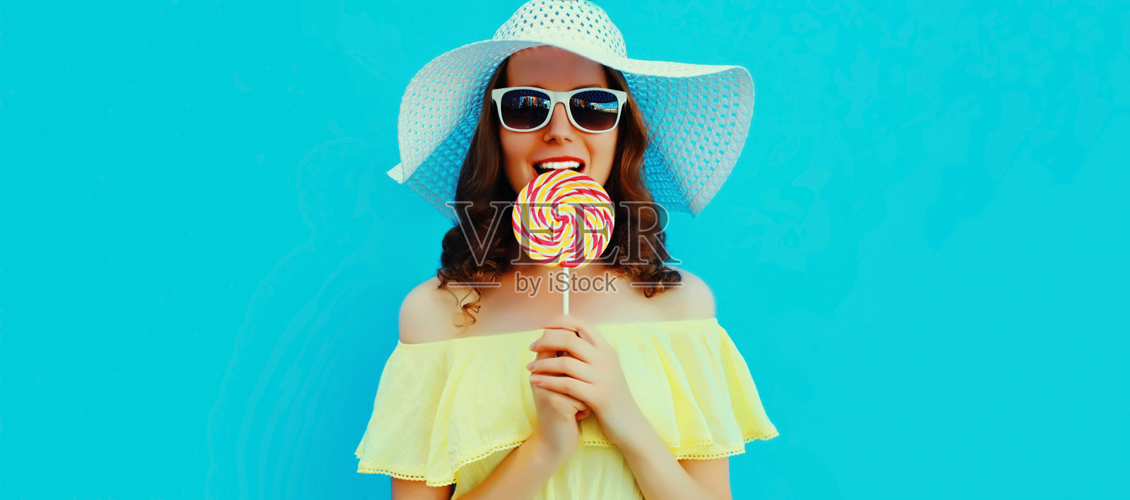 夏天快乐微笑的年轻女子，带着棒棒糖，戴着草帽，蓝色的背景照片摄影图片