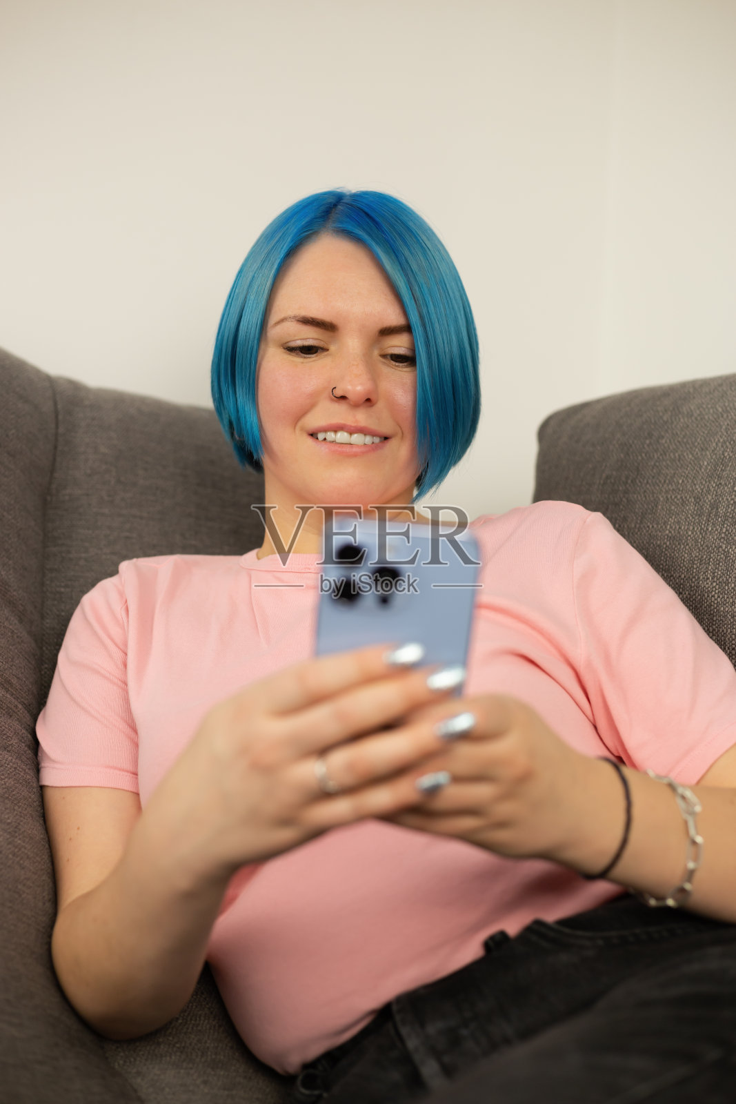 快乐的蓝头发白人女人用手机。一名年轻的成年女性，梳着染过的波波头，坐在家里的沙发上，用现代蓝色智能手机在社交媒体应用程序上打字照片摄影图片