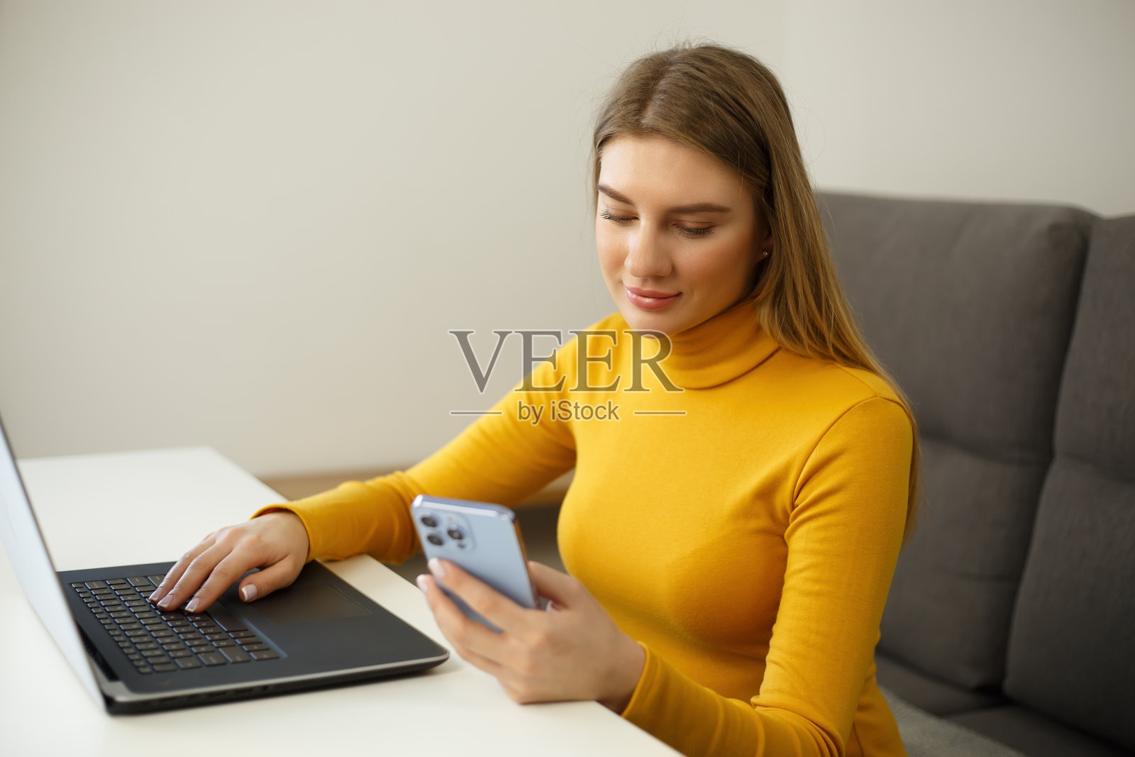 在封锁期间，年轻漂亮的金发女子使用手机和笔记本电脑进行通信和远程工作。迷人的白人女性用欢快的微笑在新智能手机上打字照片摄影图片