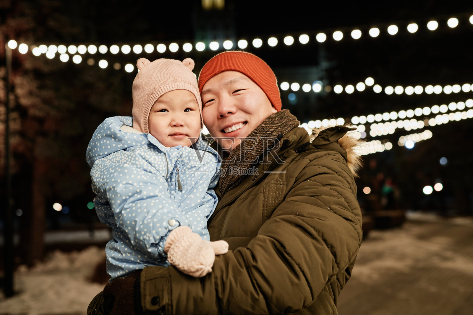 水平的中等肖像快乐的年轻亚洲人站在户外的冬季傍晚，抱着他蹒跚学步的孩子微笑着对着相机照片摄影图片