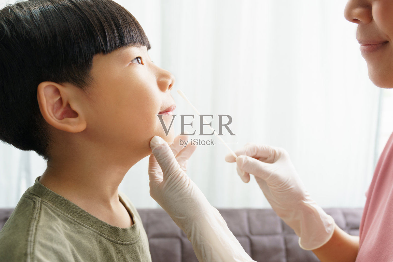 可爱的亚洲男孩戴着白色医用口罩在家里被戴着乳胶手套的母亲进行鼻拭子抗原检测。照片摄影图片