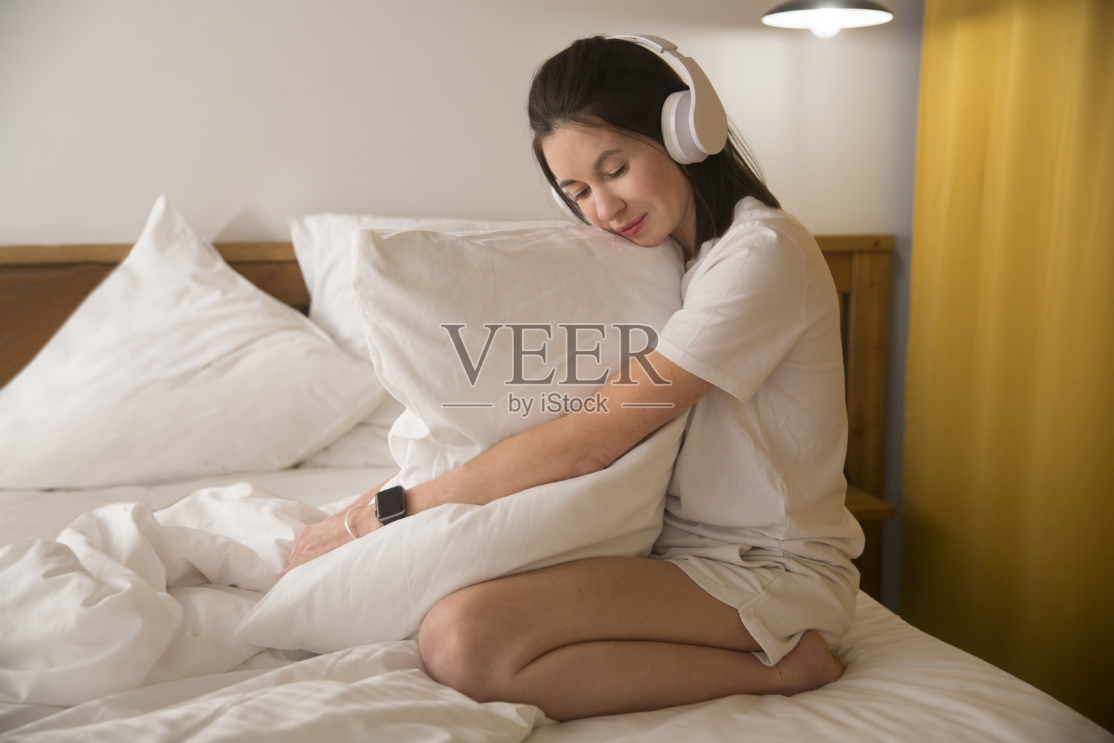 女人穿着睡衣，戴着耳机，抱着枕头，坐在卧室的床上听音乐。音频愈合。失眠。音乐和睡眠冥想照片摄影图片