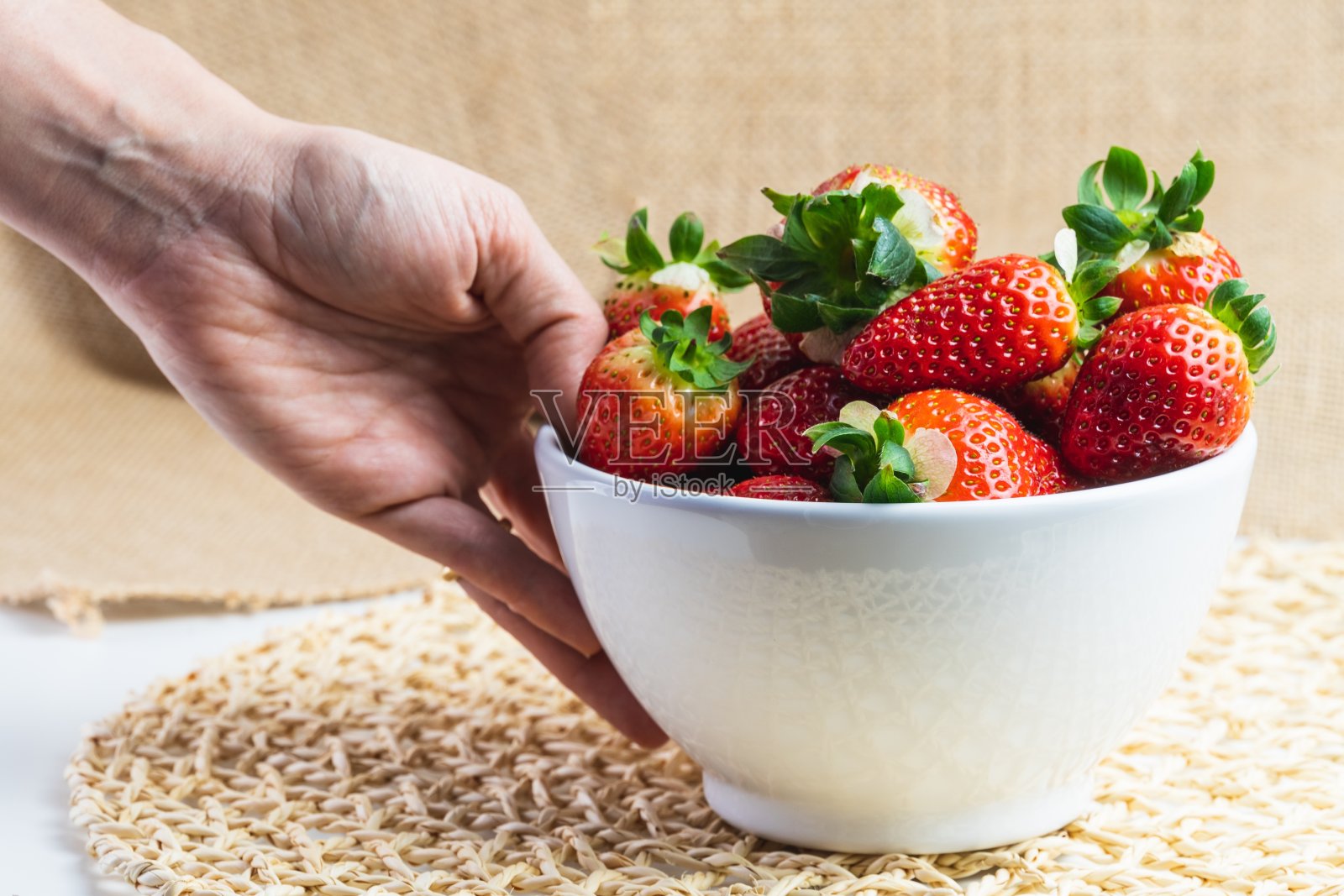 一个年轻女子的手拿起一个白色的碗，上面有几个红色的草莓，背景是乡村的。照片摄影图片