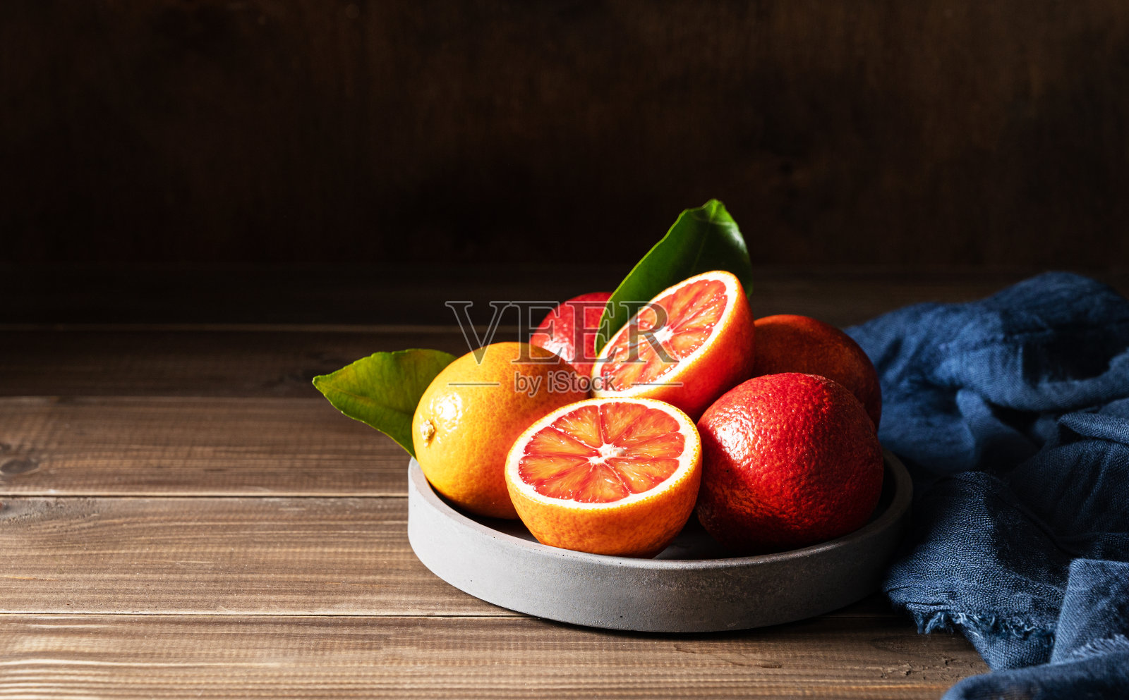多汁的红色橙子在一个混凝土碗，蓝色的餐巾在深色的木制背景。前视图和复制空间照片摄影图片