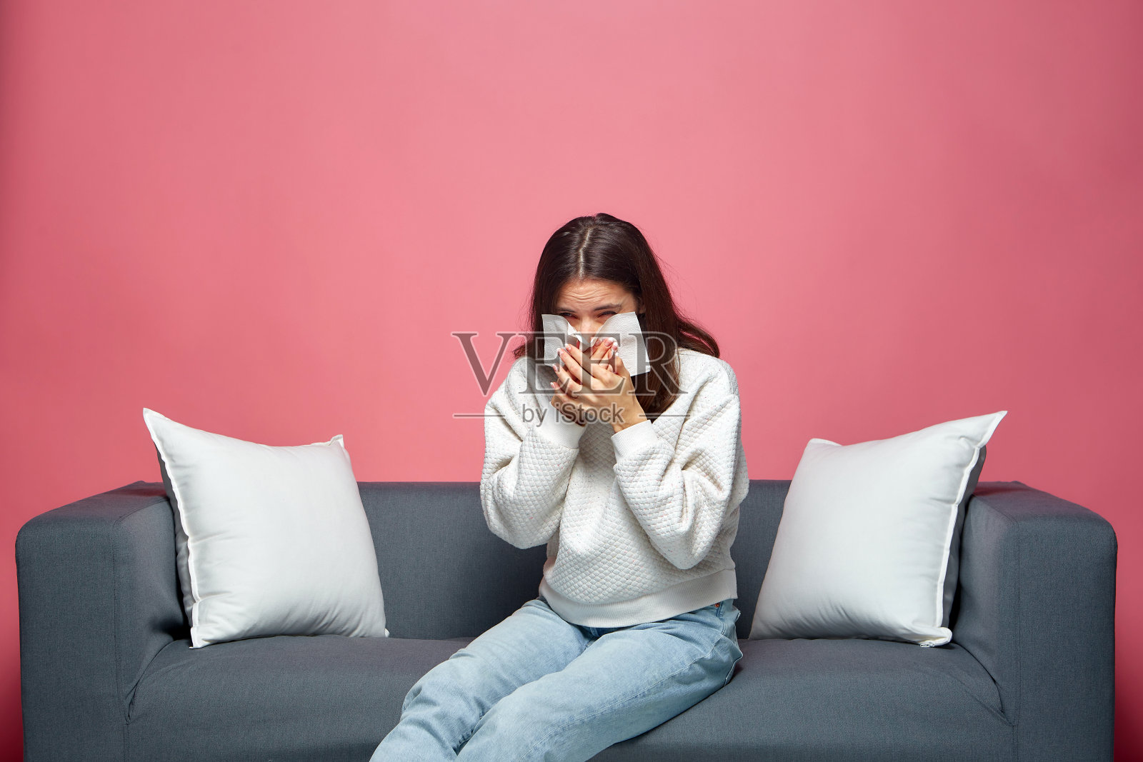 生病的年轻女孩擤鼻涕，打喷嚏，过敏，流感或感冒，坐在沙发上现代粉红色的背景照片摄影图片