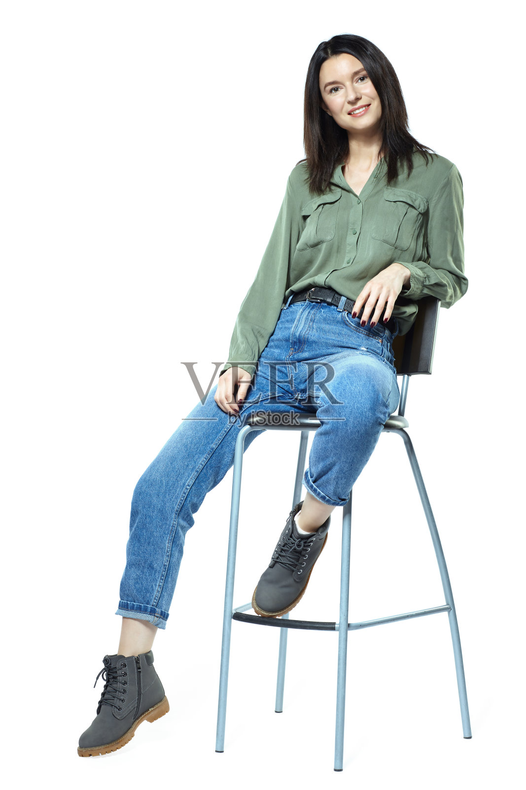 一个穿着牛仔裤、靴子和卡其色衬衫的年轻女子正坐在一把高椅上。孤立在白色的。照片摄影图片