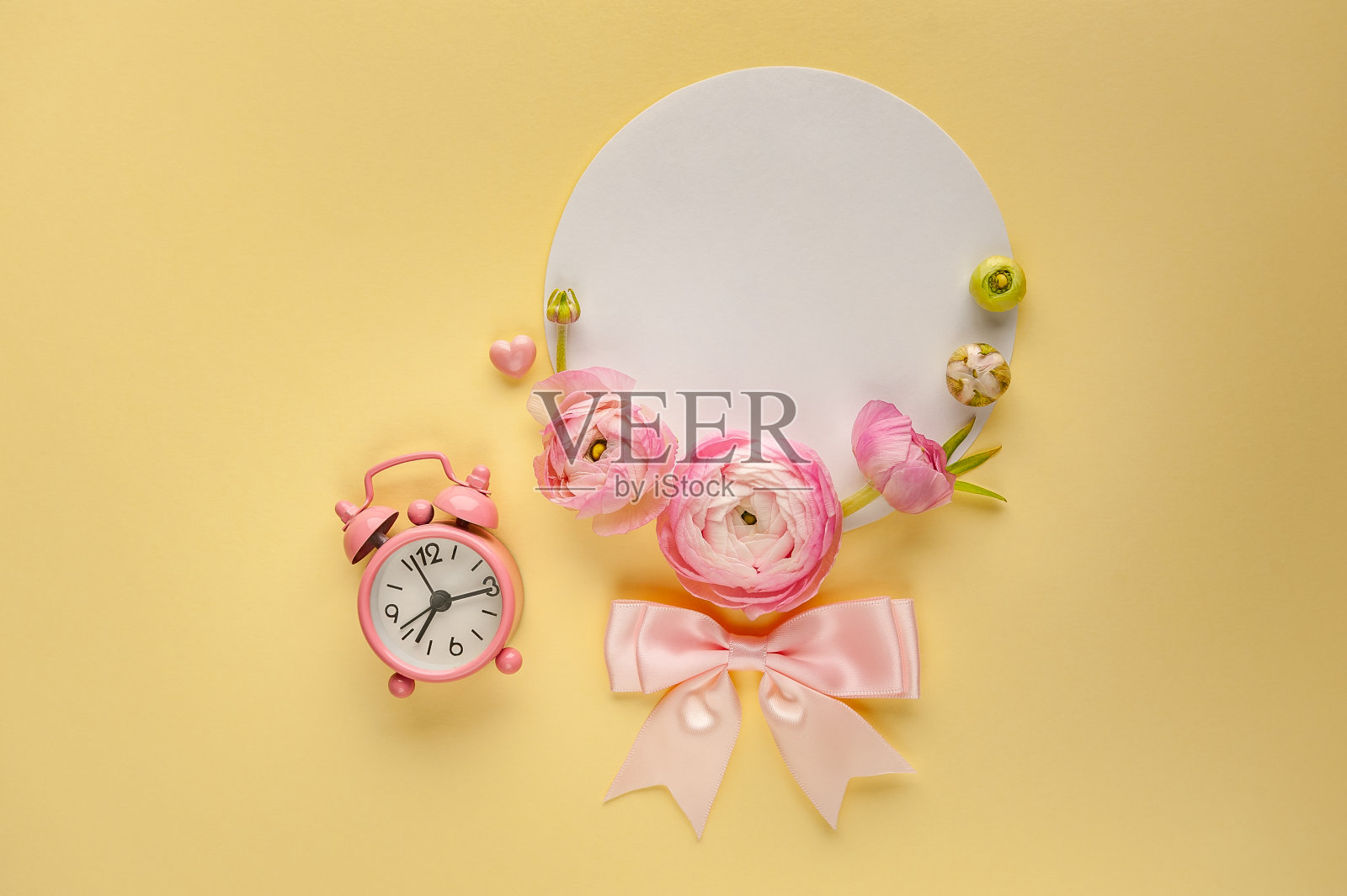 黄色背景上的空白明信片上点缀着粉红色的毛茛花和闹钟。照片摄影图片