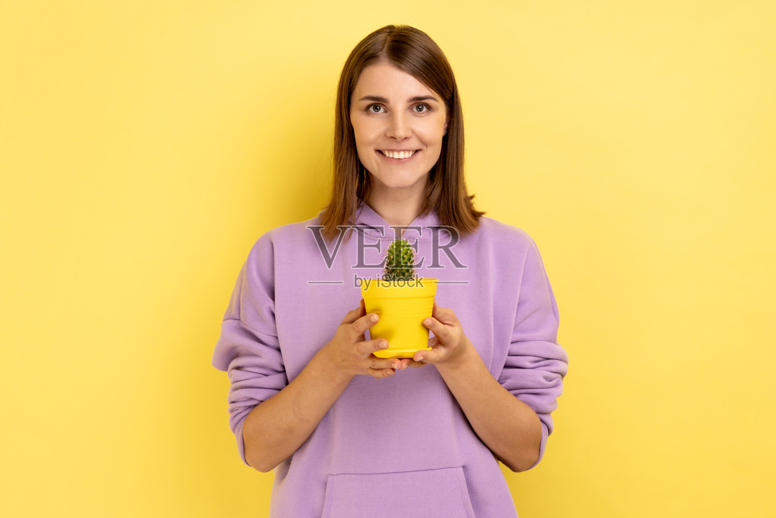 满意积极的女人拿着黄色的花盆和仙人掌，露出露齿的微笑看着镜头。照片摄影图片