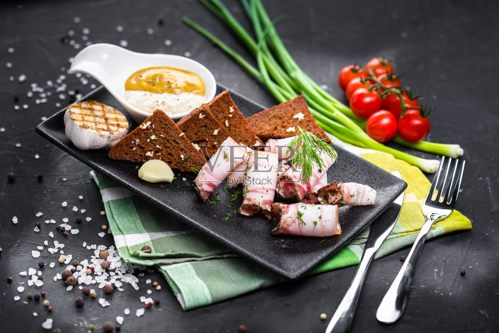 油炸黑面包丁与培根片，猪油与肉，碗与酱汁，在一个白色的盘子，蔬菜与布和木板在一个黑色孤立的背景照片摄影图片