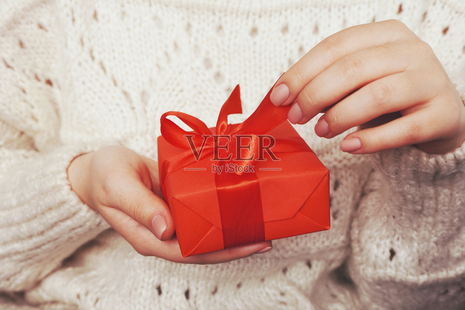 近距离拍摄的女性手拿着用红丝带包裹的礼物。穿白色或米色针织毛衣的女人。冬天的概念。情人节礼物盒子。照片摄影图片