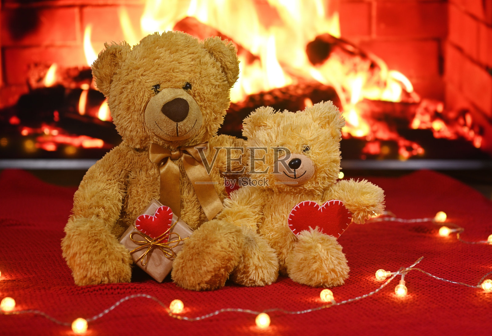 一对泰迪熊抱在一起坐着，背景是一个红色的针织格子图案和一个灯泡花环，它们的爪子拿着两个红心照片摄影图片