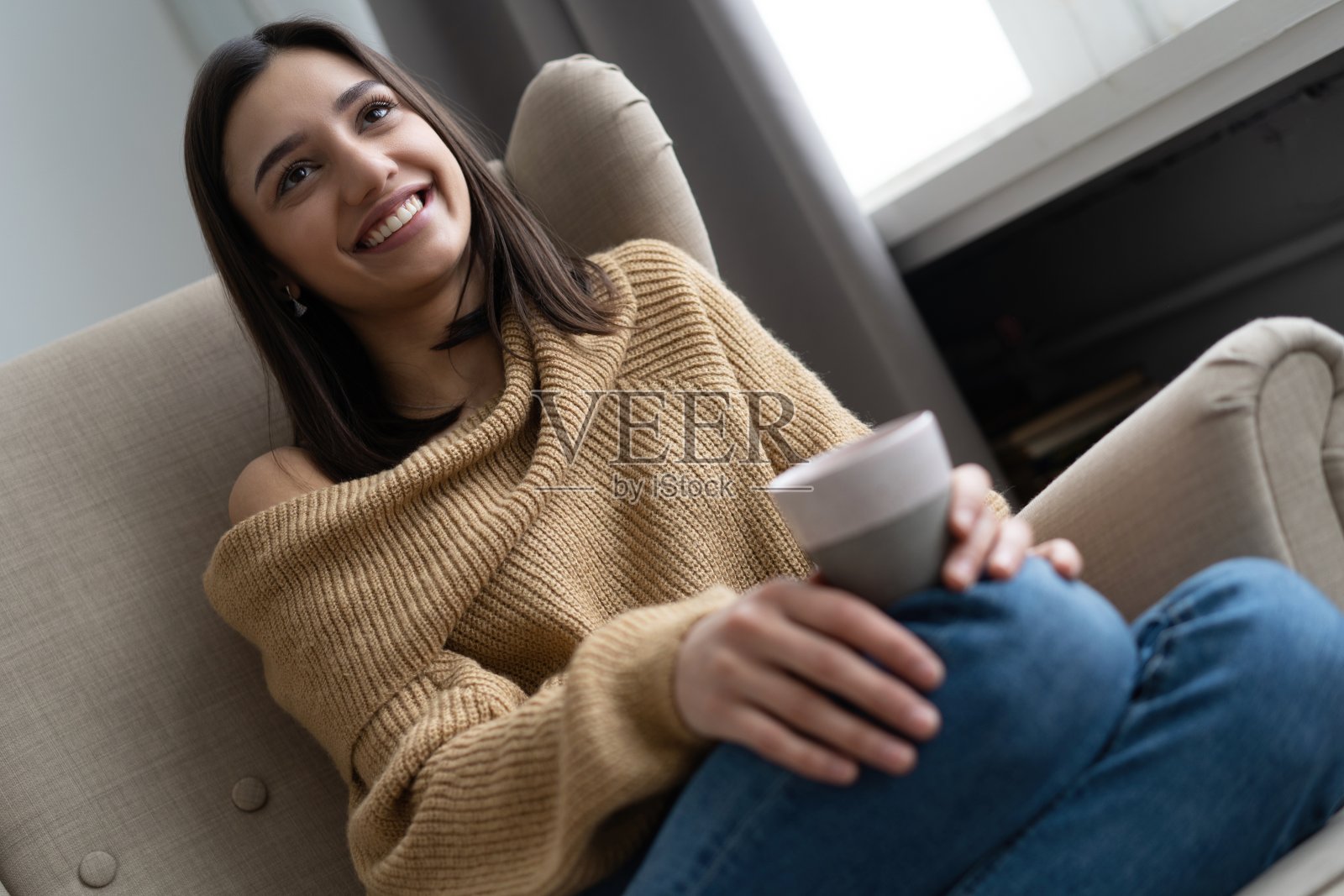 积极的年轻女士穿着休闲装坐在扶手椅上，暖手一杯热咖啡，享受着温暖的饮料照片摄影图片