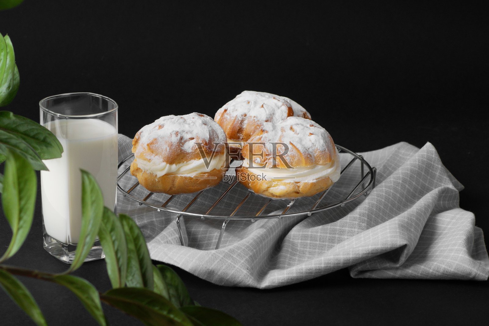 炉篦上放着新鲜出炉的奶油泡芙，炉篦下放着一条厨房毛巾。附近有一杯牛奶。这张照片是在植物的绿叶后面拍的。照片摄影图片