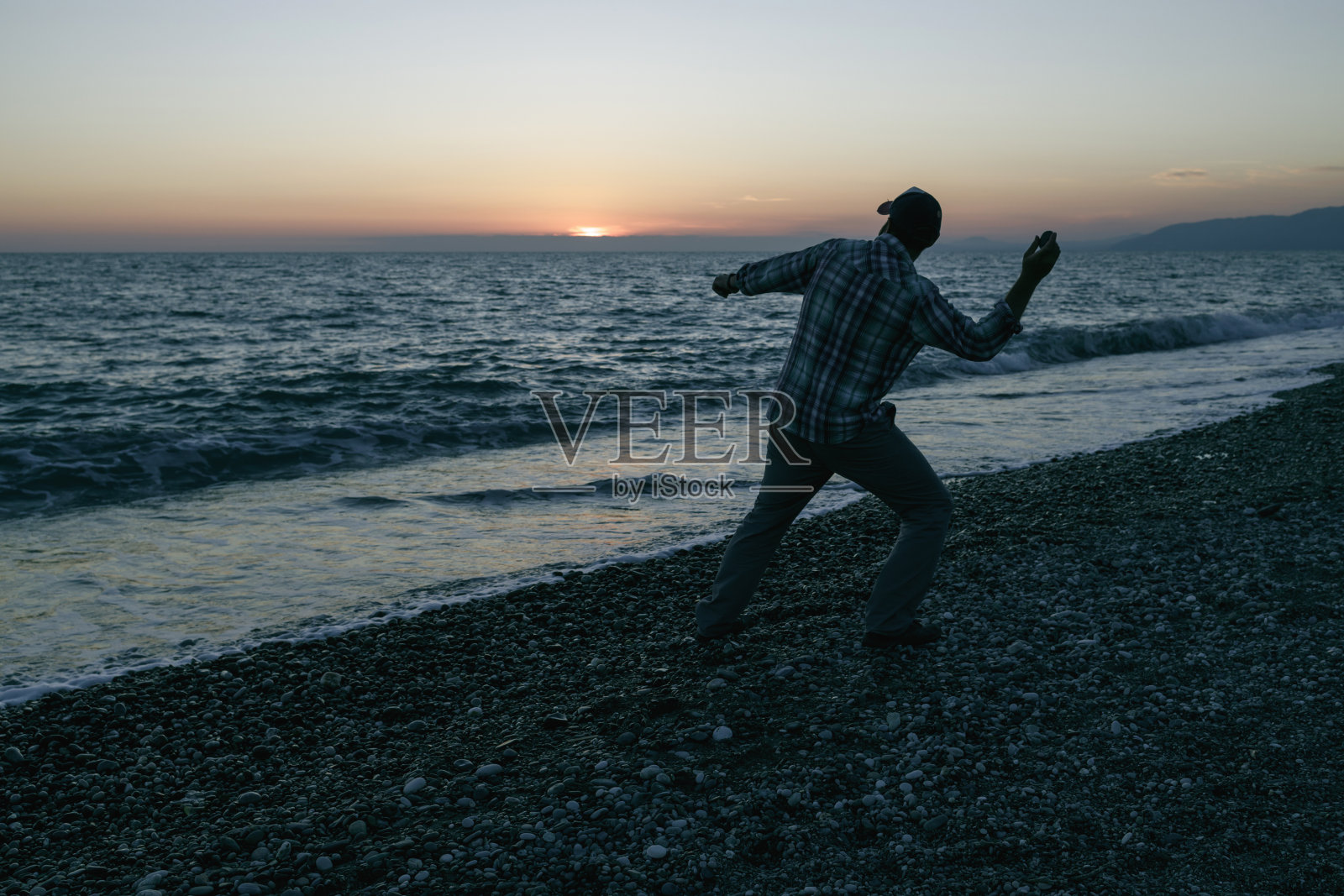 傍晚，一个穿着格子衬衫的人在海边向水中扔石头照片摄影图片