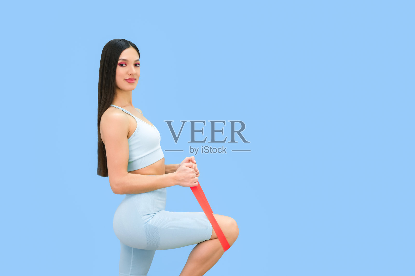 年轻漂亮的女人花时间在家里的健身房，使用健身橡皮筋，孤立在朴素的蓝色背景。照片摄影图片