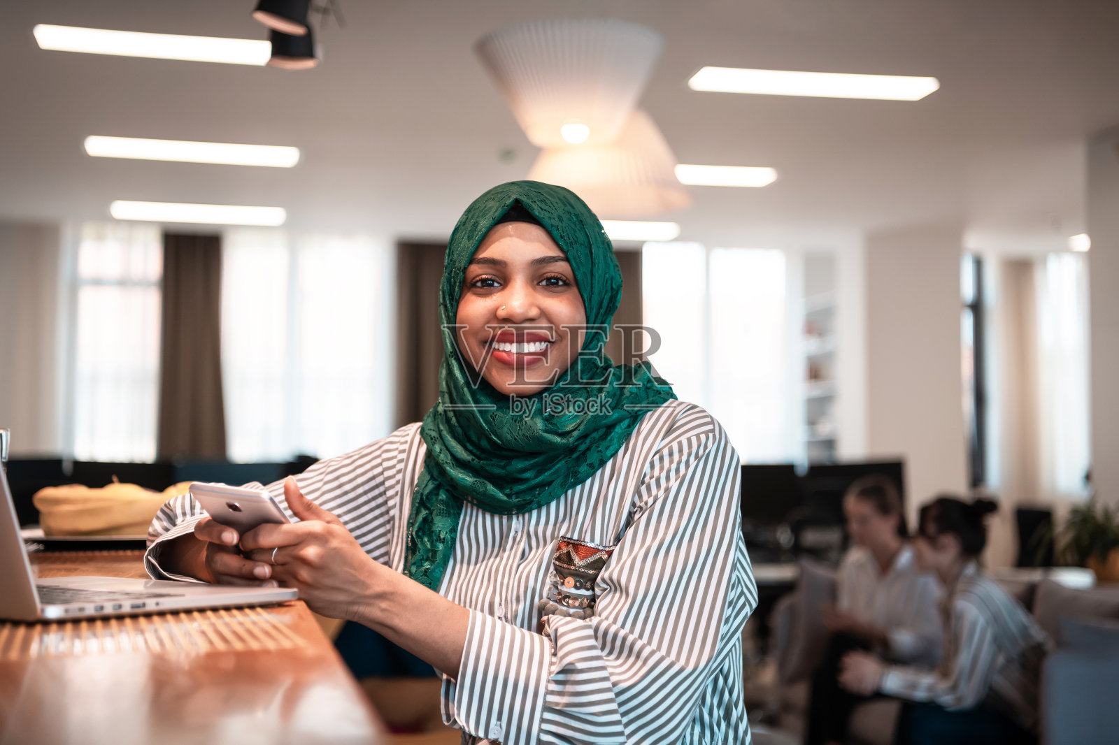 非洲穆斯林商务女性穿着绿色的头巾，在现代开放式创业办公室的休闲区使用智能手机，在笔记本电脑上工作。照片摄影图片