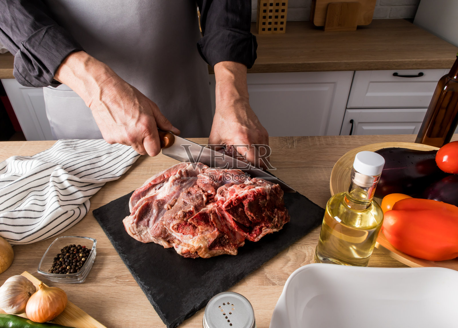 厨房桌子上的石板上，一个穿着灰色围裙的无名男子正在切一大块鲜肉。农业生态产品。照片摄影图片