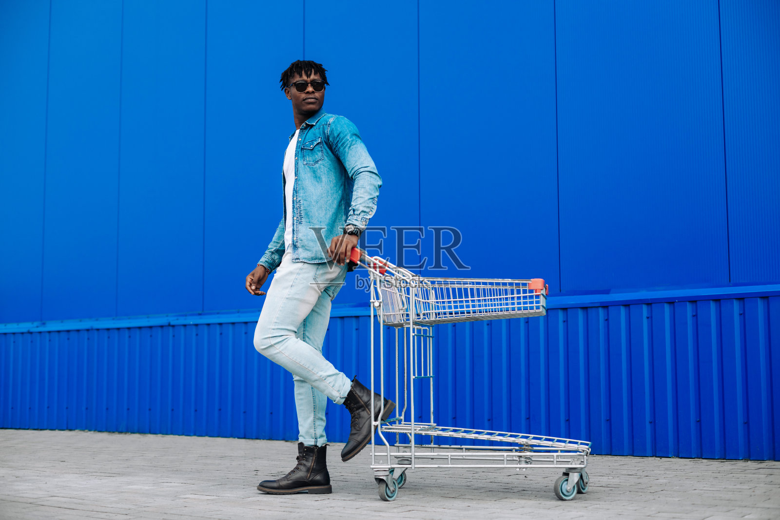 身着时尚服装，面带微笑的非裔美国人购物者，推着一辆购物车，旁边是户外的蓝色墙壁照片摄影图片