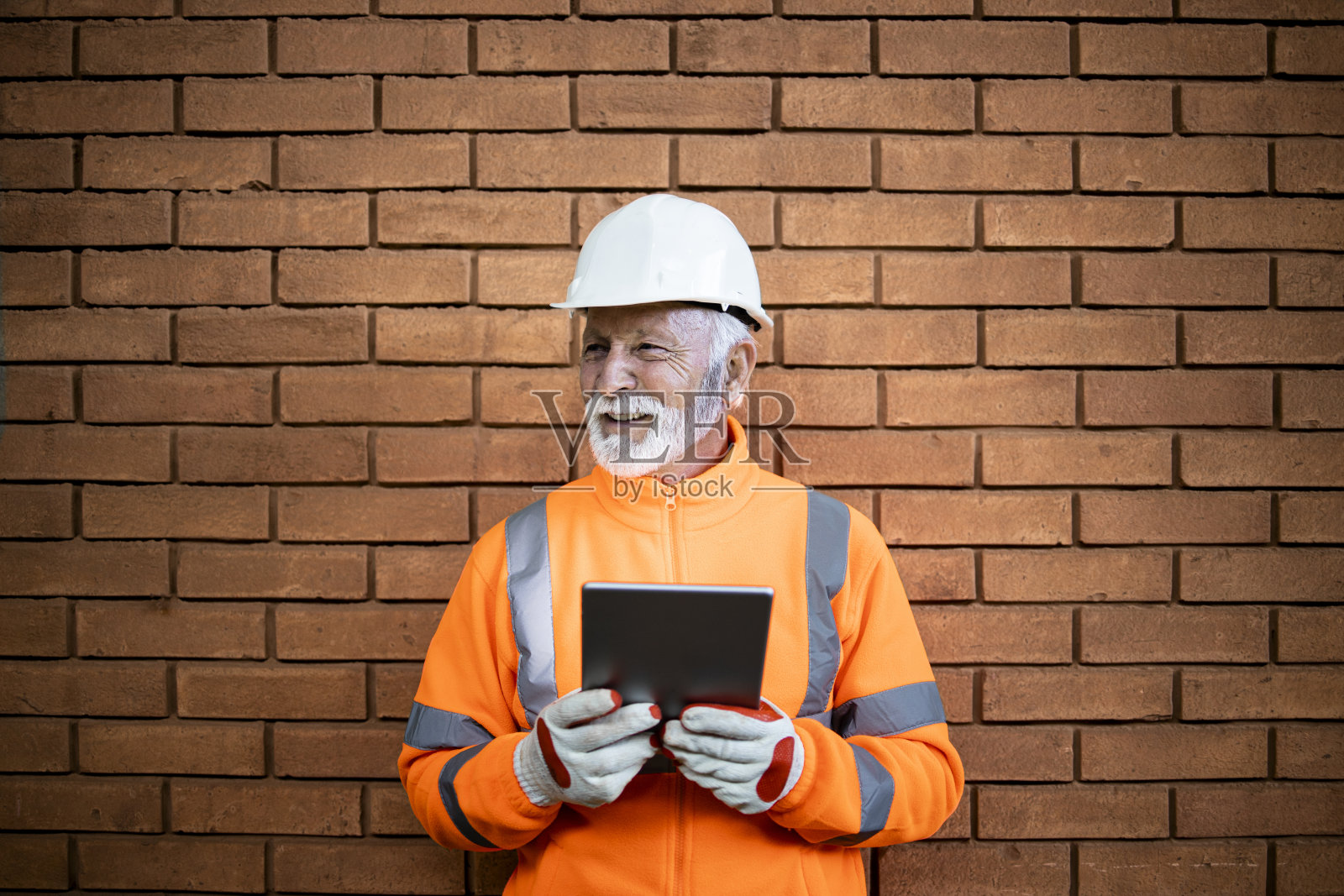 一名经验丰富的建筑工人戴着安全帽和安全设备站在工地的砖墙旁。照片摄影图片