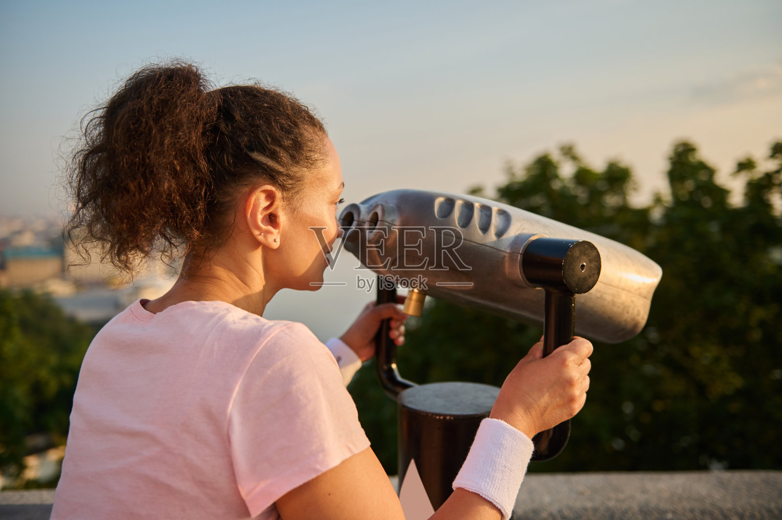 一个漂亮女人用望远镜看城市的后视图。穿着粉色t恤的女运动员正通过静止的城市望远镜观看美丽的绿色自然和河流背景照片摄影图片