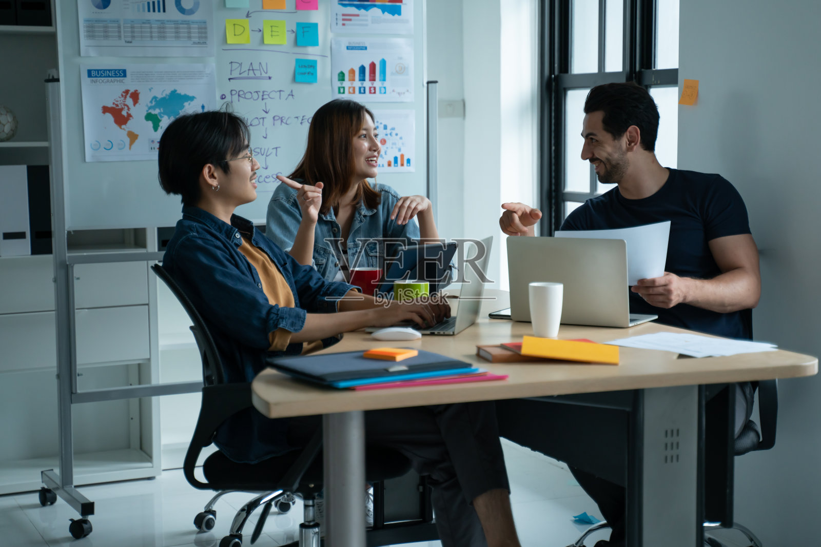 一群穿着休闲服装的年轻商务人士坐在办公桌前，与坐在后面的同事一起工作和交流。商务人士会面概念。照片摄影图片