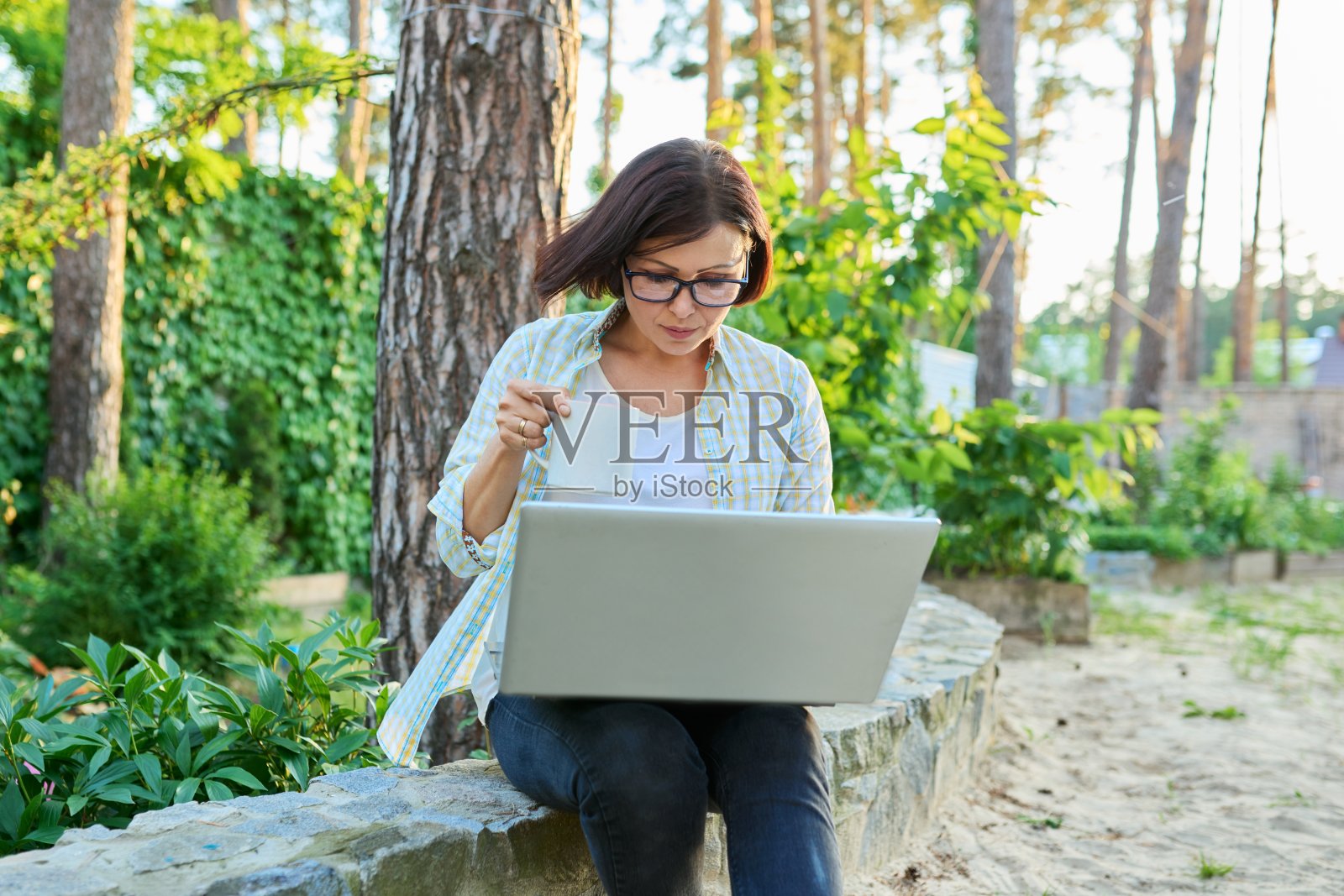 严肃的中年妇女与笔记本电脑杯休息工作在后院。照片摄影图片