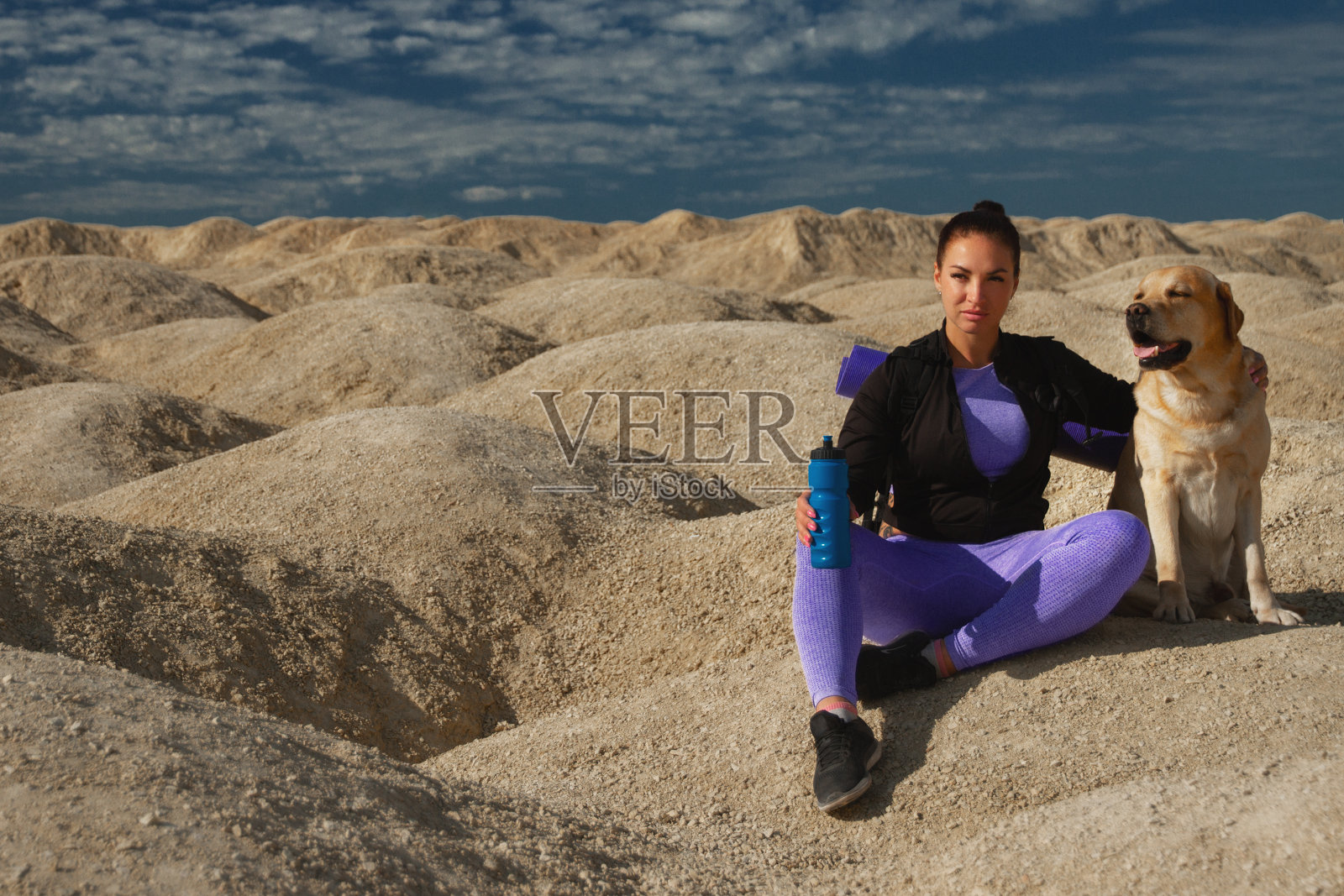 在阳光明媚的日子里，一个女人背着运动蓝色的双肩包坐在黄色的沙丘上，和一只拉布拉多猎犬一起望向远方照片摄影图片