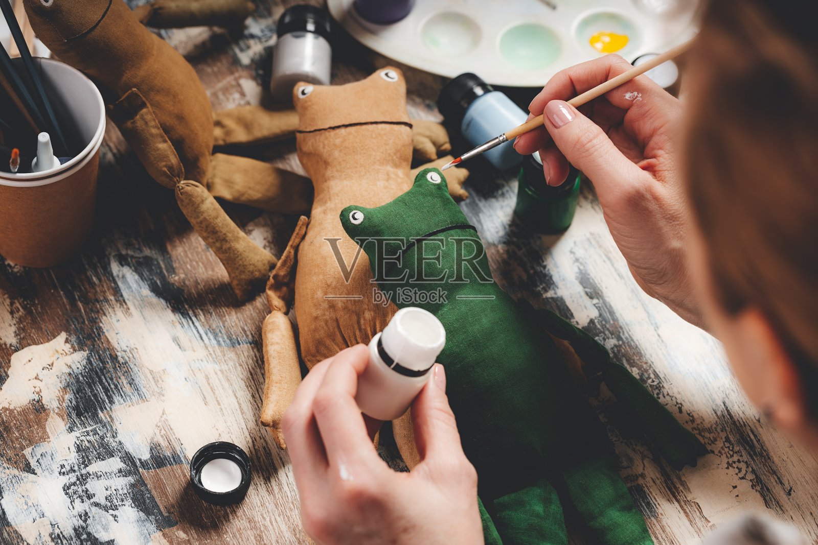 手工制作的软娃娃青蛙。DIY制作的礼物。人类手握青蛙软玩具。刺绣配件在灰色的背景。手工制作的装饰房子。制作老式软玩具的过程。家庭作坊。照片摄影图片