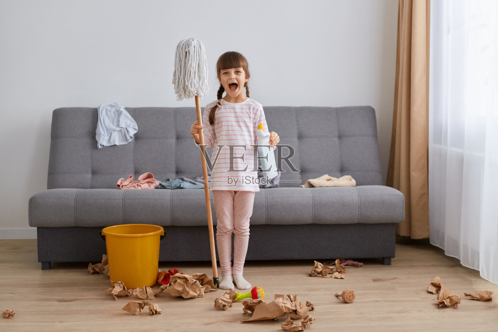 可爱的黑发小女孩，穿着休闲的衣服，正在打扫房子，手里拿着拖把站着，张大嘴巴看着镜头，需要洗地板。照片摄影图片