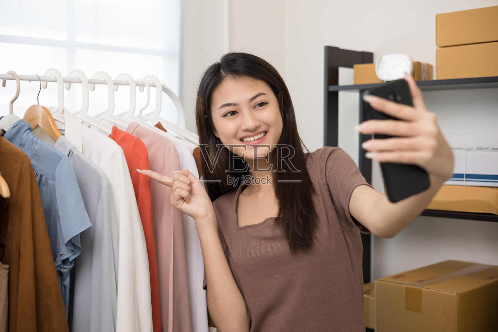 年轻的亚洲女性企业主在时尚商店使用智能手机直播销售时尚服装。博主评论在社交媒体上展示的服装。网上购物的概念。照片摄影图片
