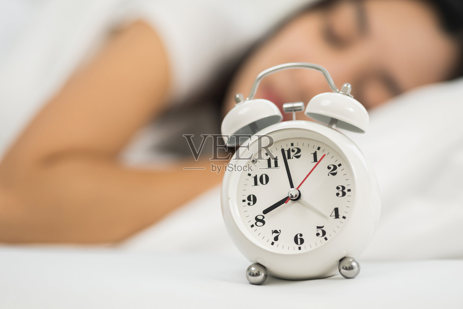 美丽的女人早上在床上被闹钟叫醒。年轻的亚洲女性不想醒来，也不喜欢闹钟的声音。照片摄影图片