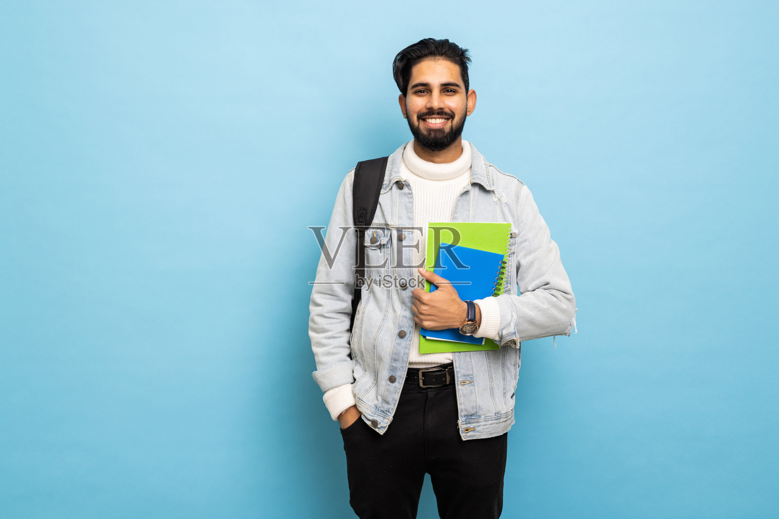 英俊的年轻印度男子背着双肩包，拿着蓝色背景的书照片摄影图片