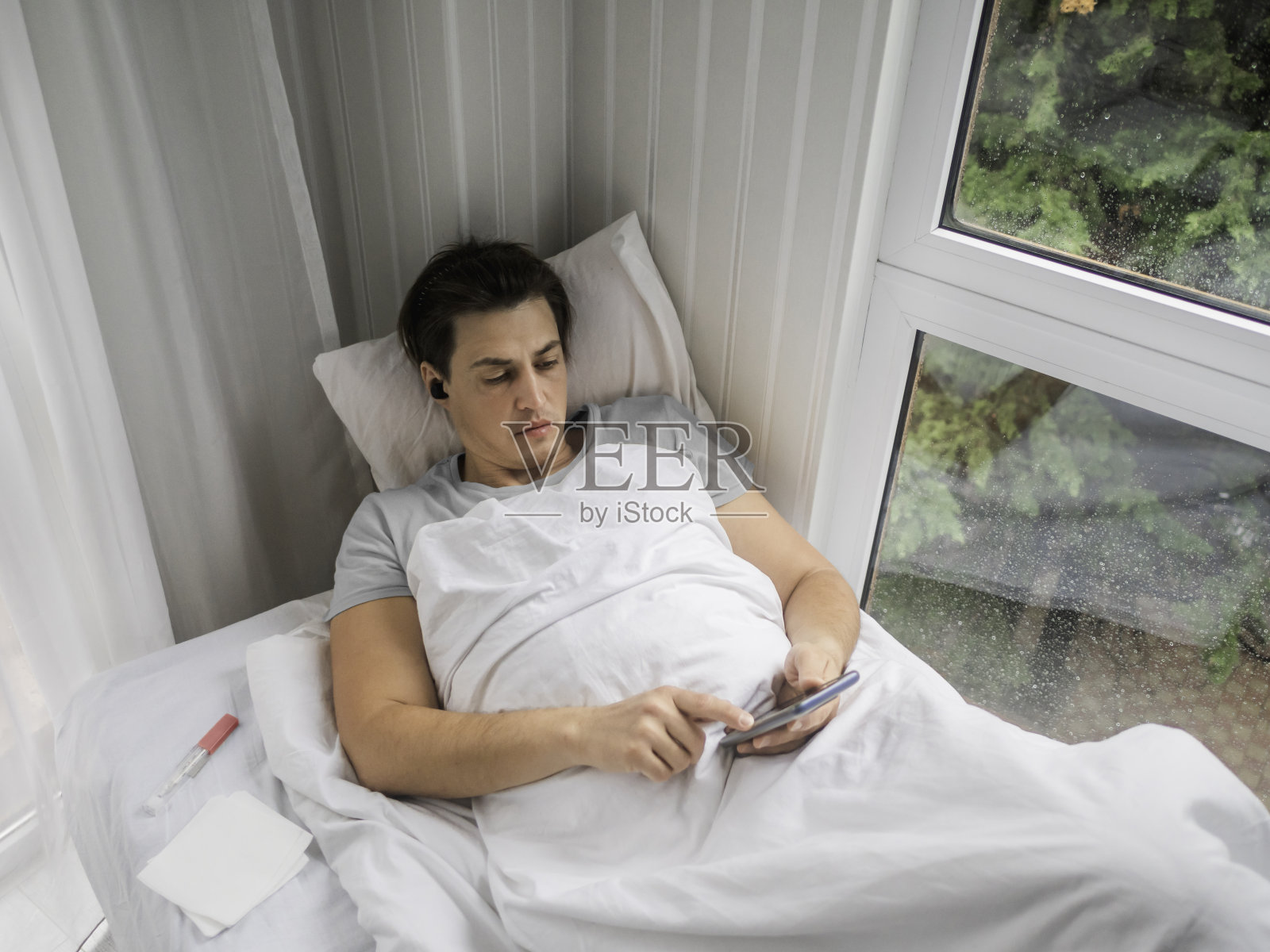 病人拿着体温计躺在床上。一名男子正在网上搜索疾病症状或给他的医生发短信。在暂时残疾的情况下使用无线技术。照片摄影图片