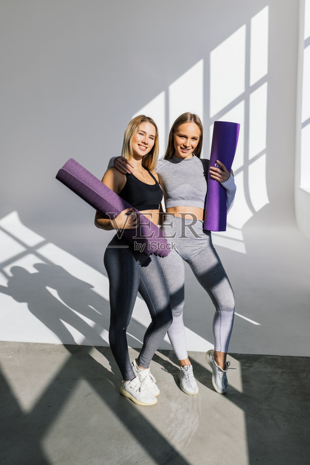 两个穿着运动服的年轻女性朋友在瑜伽垫旁散步，在健身房的瑜伽休息时间聊天和玩乐照片摄影图片