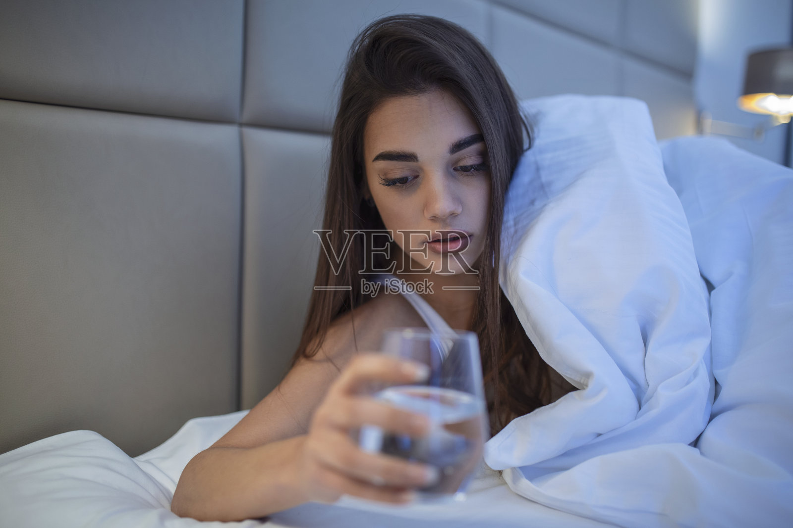 年轻女子晚上在床上喝一杯水。女人睡前喝一杯水，她正躺在床上照片摄影图片