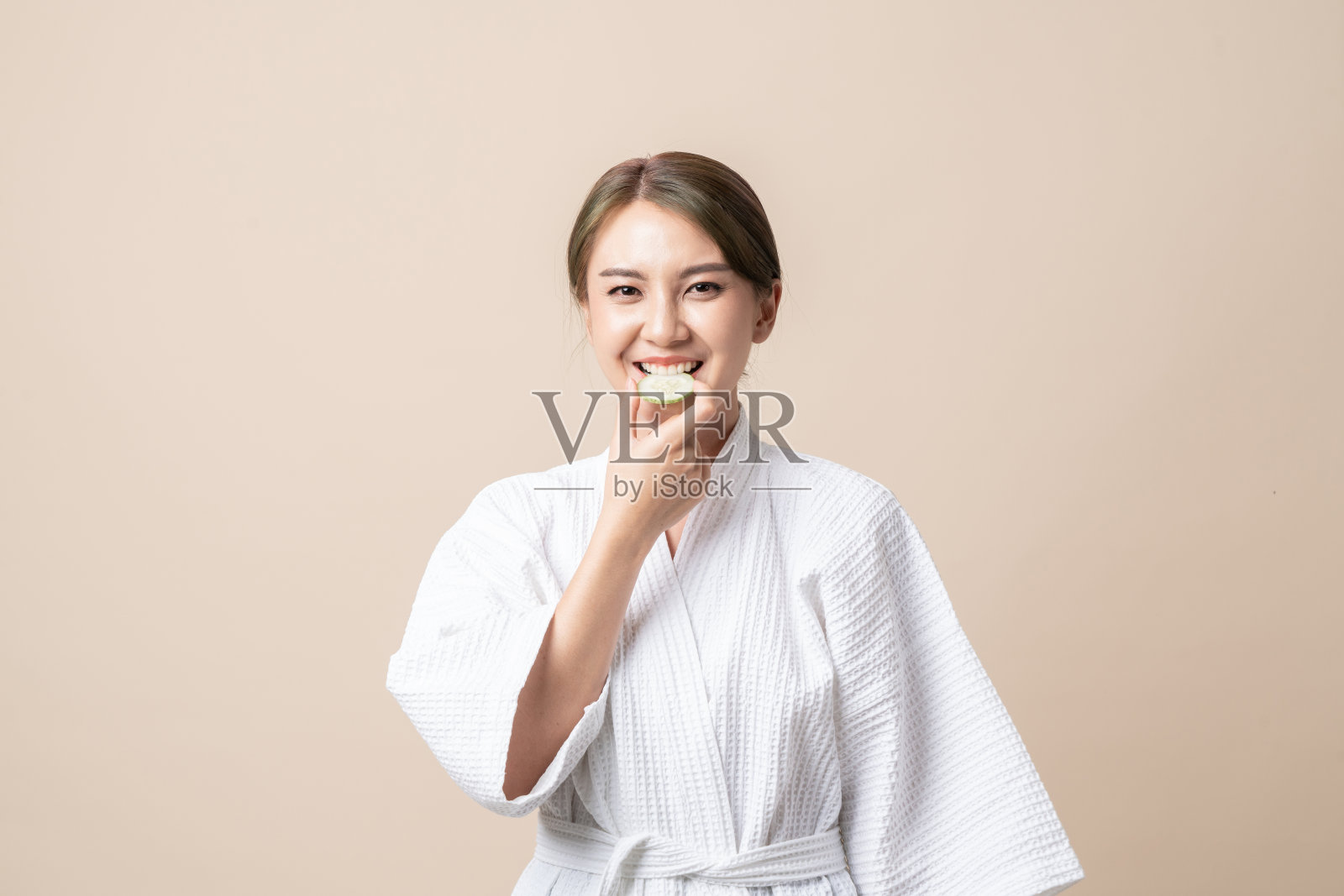 美丽的年轻亚洲妇女在浴衣与持有黄瓜片面部面具在棕色孤立的背景。照片摄影图片