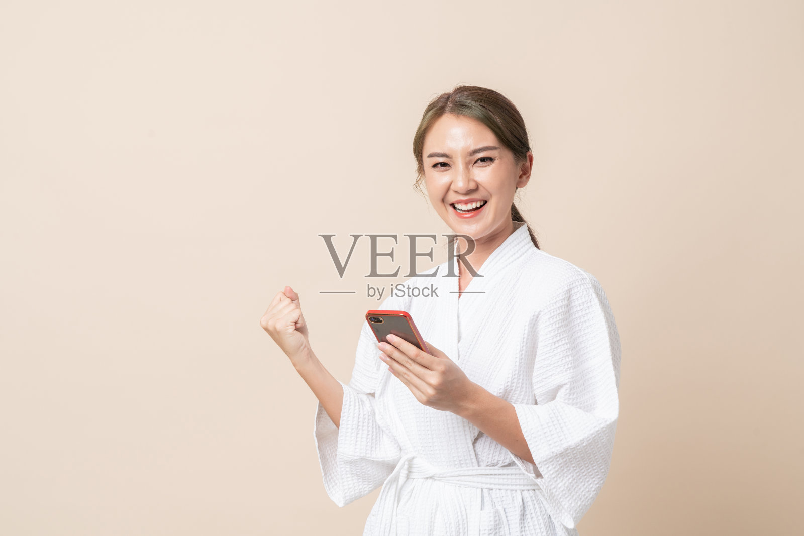 肖像美丽的年轻亚洲妇女幸福的微笑在浴袍使用智能手机在棕色孤立的工作室背景。照片摄影图片