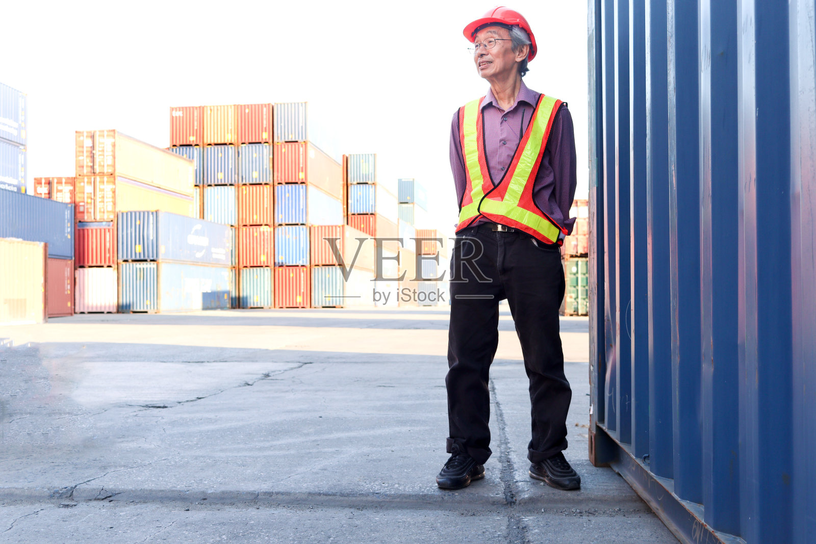 照片中是一位年长的亚洲工人工程师，身穿安全背心，头戴安全帽，站在物流运输集装箱堆场。照片摄影图片