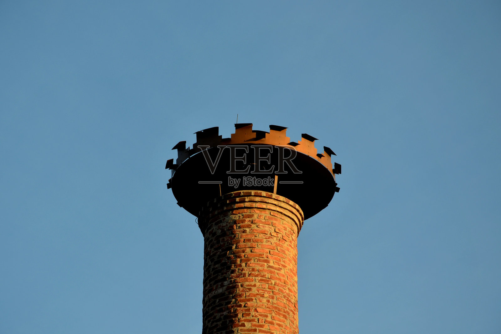 啤酒厂历史悠久的砖砌烟囱上有一个用于白鹳筑巢的金属齿状结构。褐色的砖烟囱。锡冠上方的洞照片摄影图片