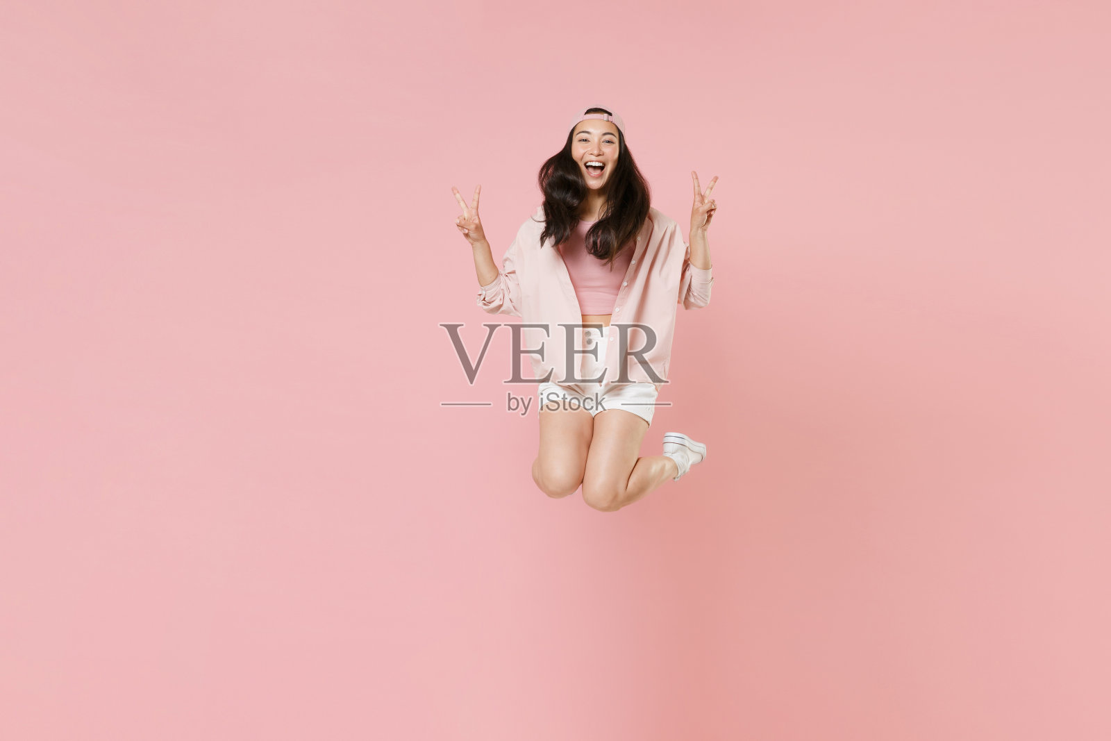 快乐的年轻亚洲女孩在休闲服装的全长肖像，帽子孤立在柔和的粉红色背景工作室肖像。人们生活方式的概念。模拟复制空间。跳起来，露出胜利的手势。照片摄影图片